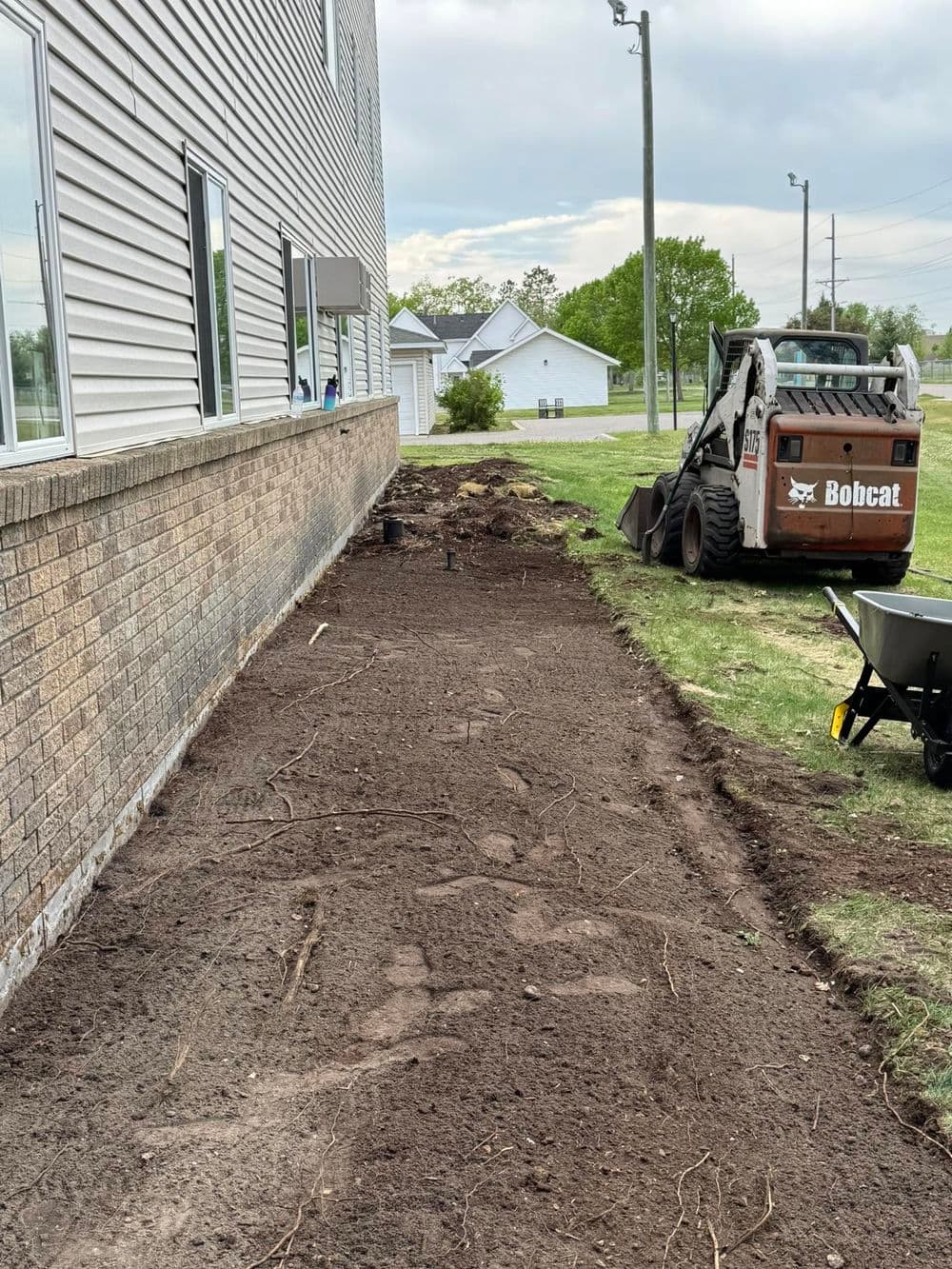 Landscape preparation in progress beside a building, with a Bobcat skid steer and tools.