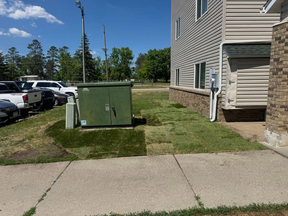 Green utility box next to a building with freshly cut grass and parked vehicles nearby.